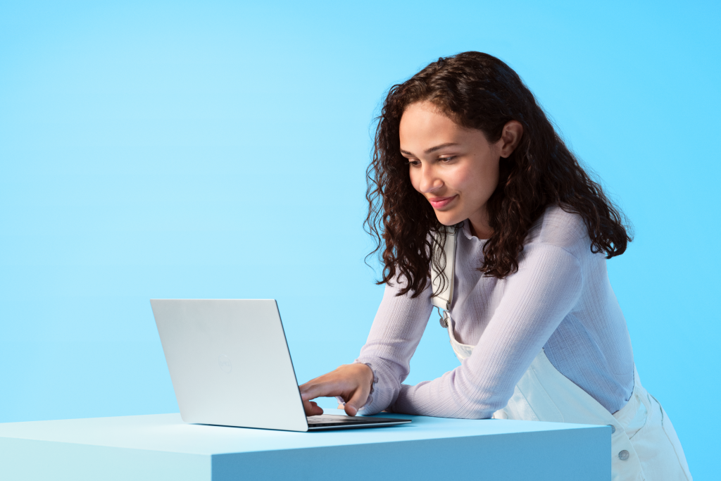 Frau mit braunen Locken und weißer Latzhose an einem Laptop vor blauem Hintergrund