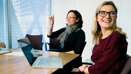 Two smiling women at a conference table, one with a laptop in front of her and the other raising her hand to speak