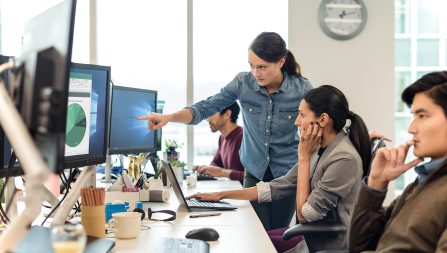 Female IT employee in conversation with female coworker. The IT employee is standing over workstation, pointing to desktop monitor, and providing explanation to seated coworker who is using a Dell laptop. Two male coworkers are also sitting at the workstation looking at their desktop monitors