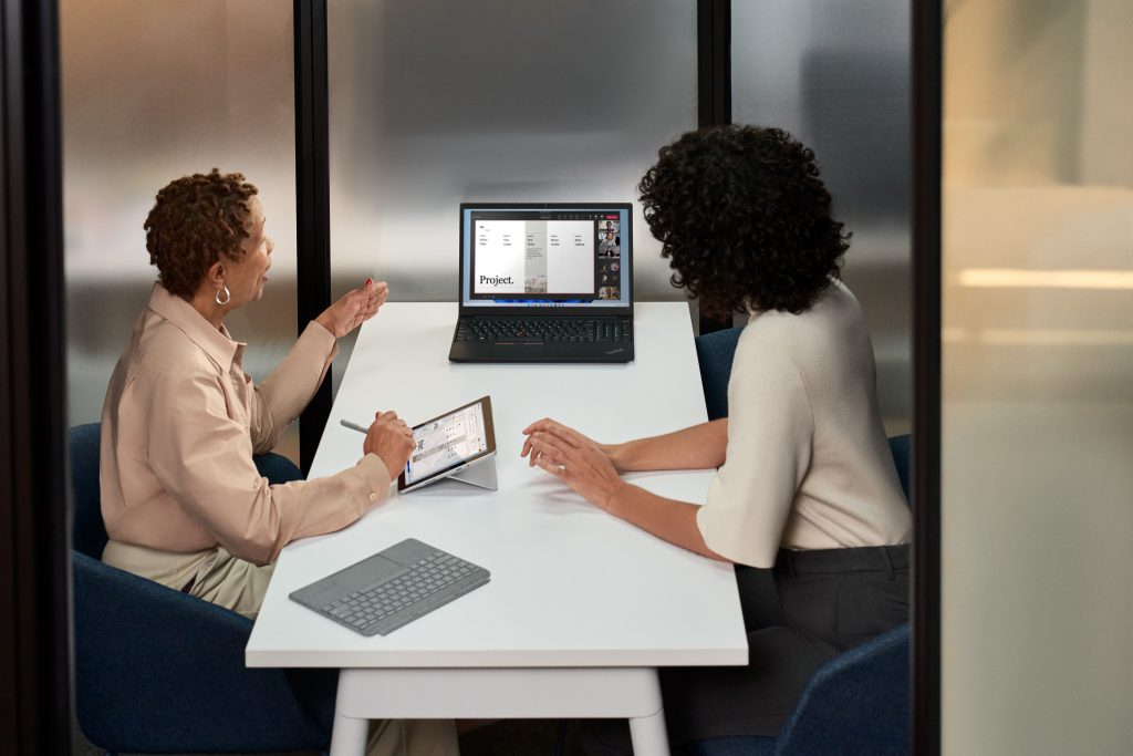 Two women sitting in a room staring a computer desktop and introducing a project to a group of people over Microsoft Teams.