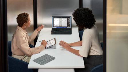 Two women sitting in a room staring a computer desktop and introducing a project to a group of people over Microsoft Teams.