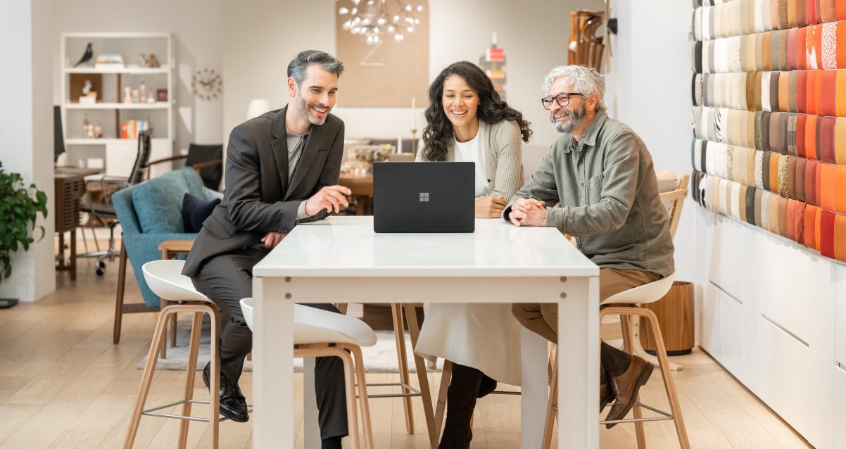 3 people sitting at a desk and looking into a computer while smiling and being communicative.