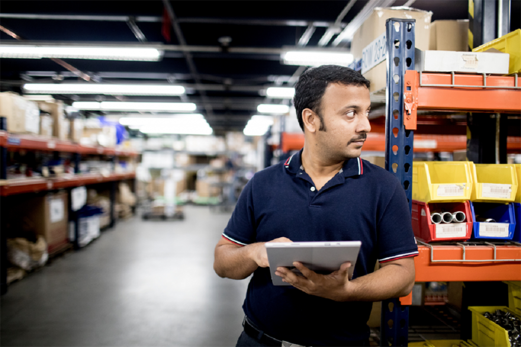 Man standing among warehouse shelves holding a tablet.