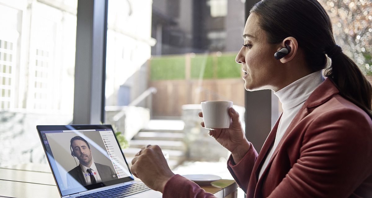Woman in office wearing an earpiece, holding a cup, and conducting a video call with a male colleague