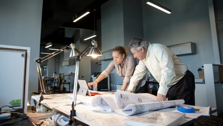 Two engineers looking over blueprints and a laptop screen