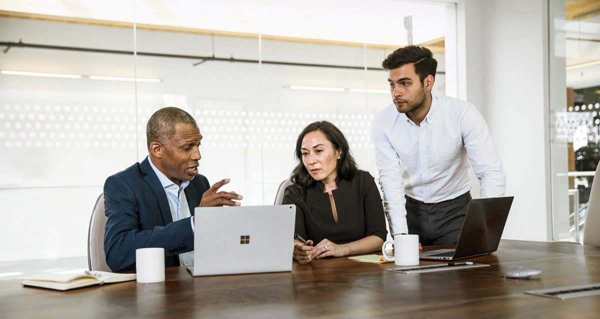 Three coworkers at a conference table chat while looking at a laptop