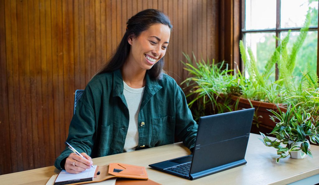 Women looks at laptop and smiles while she jots down notes