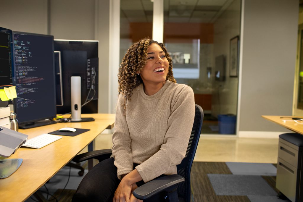 A worker smiling at her desk with computer code on the monitor